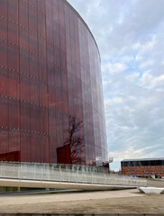 Modern building with a curved red glass facade reflecting the sky and surroundings, featuring a...