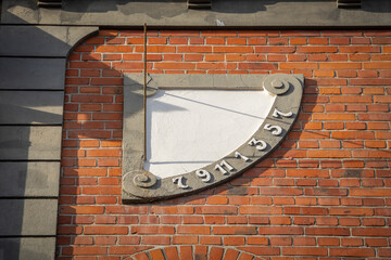 A sundial on a red brick wall with a gray plaster frame shows the hours from 7 to 5, illuminated by the low sun