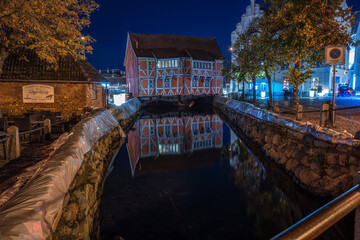Night scene with an illuminated half-timbered house on the canal, calm water and reflection, surrounded by cobblestones and autumn trees
