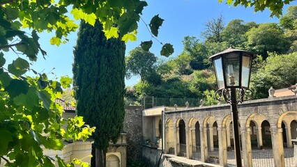 Serene architecture: Sunlight filters through lush foliage, illuminating the intricate stonework and classical architecture of a historic structure with a vintage street lamp.