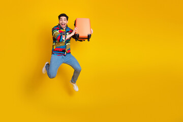 Happy young man in a colorful shirt jumps mid air holding an orange suitcase against a bright yellow background promoting travel and fun lifestyle