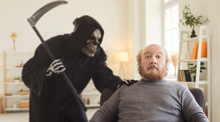 Retired old senior man sitting in armchair at home with shocked scared face expression feels sudden breath of death as Grim Reaper in hooded dark cloak approaches from behind. Death concept