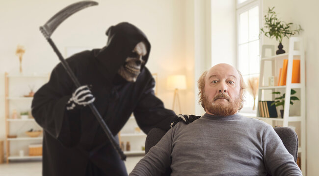Fototapeta Retired old senior man sitting in armchair at home with shocked scared face expression feels sudden breath of death as Grim Reaper in hooded dark cloak approaches from behind. Death concept