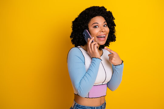 Fototapeta Cheerful young woman pointing excitedly while on the phone against a vibrant yellow background, showcasing joy, style, and communication