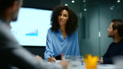 Business meeting in a modern glass-walled conference room, diverse team of professionals sitting around a table while a presenter stands and points at a large digital  - Powered by Adobe