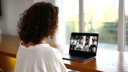 woman participating in an online business meeting from home, rear view showing her at a wooden desk with a laptop displaying a video conference of 12 diverse colleagues, natural - Powered by Adobe