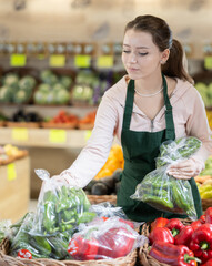 Young woman saleswoman is standing near the counter in a vegetable store and putting green peas in bags. Stalls at the market with full baskets of vegetables and fruits