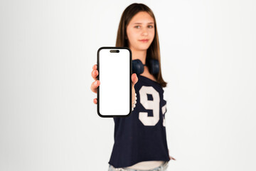 Fototapeta premium Teenage girl with long hair holds a smartphone with a blank white screen, wearing a dark shirt and headphones, isolated on white. Suitable for technology, app promotion, advertising, and digital