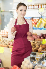 Saleswoman in a red apron holds potatoes against the background of a display case with vegetables. Farm market employee lays vegetables on the counter