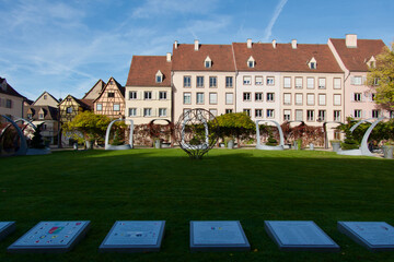 Fototapeta premium colmar Charming European Square with Historic Half-Timbered Buildings Featuring Modern Art Sculptures Amidst Greenery Under Clear Blue Sky