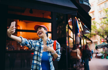 Cheerful Asian woman laughs while recording a selfie video outdoors, holding phone with one hand and touching chest with the other, radiating emotion, connection and digital authenticity.