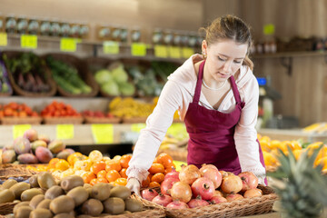 Saleswoman in an apron stands near the counter in a fruit shop and holds a basket of ripe pomegranates in hands. Employee of the vegetable department of a supermarket puts a box of fruit