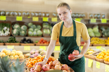 Young woman works as a seller in a fruit shop, she carefully puts a pomegranate on the counter. Supermarket worker checks the quality of products