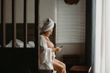 Mature caucasian female in bathrobe using phone by window in cozy room