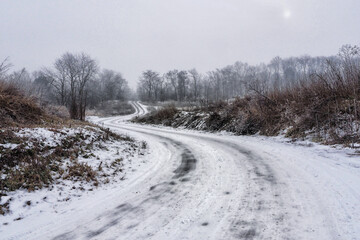 Snow covered country road curving through frosty fields. Winter landscape with misty trees and soft light.