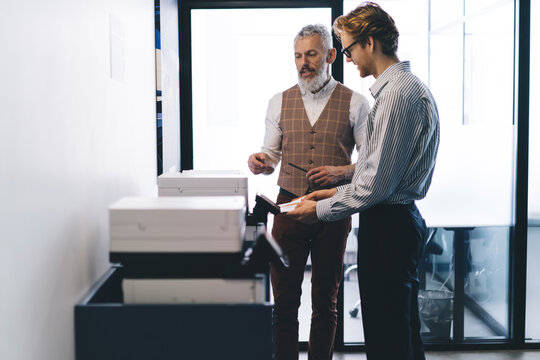 Senior and junior businessmen operate office printer together, showcasing teamwork, process navigation, and supportive environment within modern digital workplace.