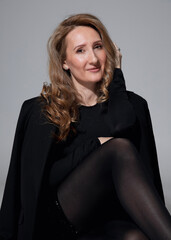 Fototapeta premium Front image of an elegant slim woman with makeup, dressed in black clothes, looking at the camera, sits down on the floor with crossing legs. 