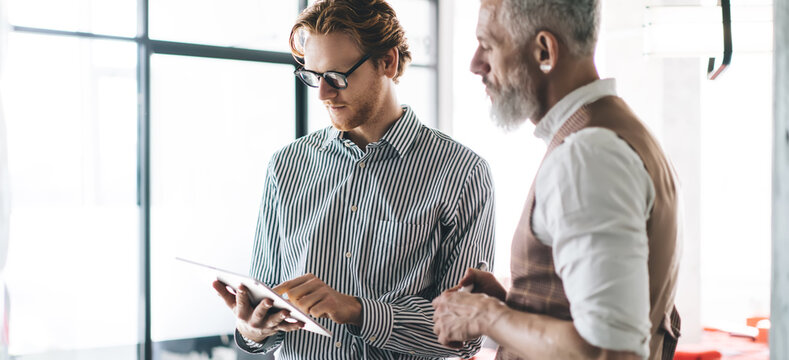 Young businessman scrolls on tablet while older colleague watches and listens. Moment captures hands-on tech usage, cross-generational communication, and flexible knowledge transfer.