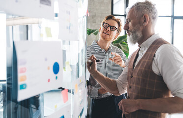 Senior mentor writes on glass wall as younger colleague watches attentively. Scene captures...