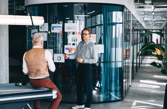 Young businessman presents data to older partner near glass wall with charts. Symbolizes visual leadership, startup communication, and real-time teamwork with tech and infographics.