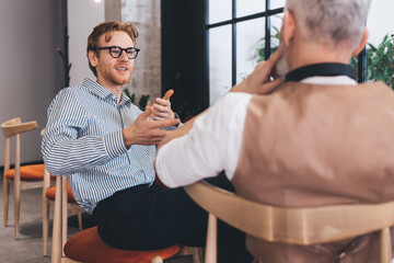 Young male professional explains with expressive gestures in conversation with older colleague. Moment captures idea pitching, intergenerational dialogue, and flexible leadership.