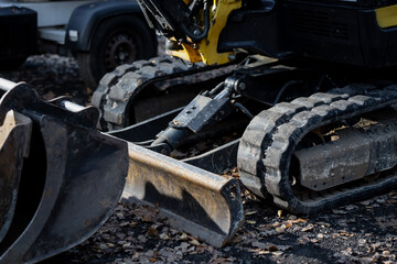 Compact excavator with rubber tracks and steel grading blade parked on outdoor worksite covered with dry leaves showing construction machinery details and hydraulic mechanism design