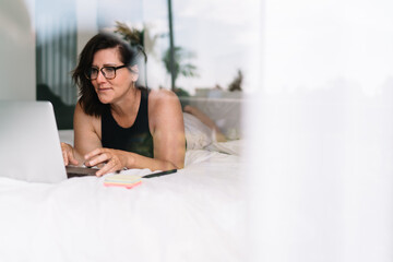 Serious middle-aged freelancer deeply focused on laptop screen through window, symbolizing...