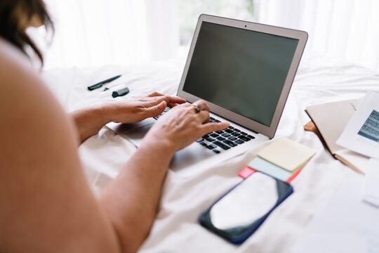 Close-up of female freelancer typing on laptop while working remotely from bed, surrounded by smartphone, sticky notes, and essential digital tools for organized tech-focused workflow.