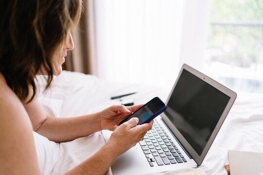Woman freelancer checking smartphone beside laptop, combining attention, tech tools and relaxed pace to handle remote business tasks efficiently from a digital home setup.