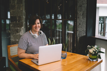 Freelancer smiling at camera while working on laptop indoors, headphones around neck, representing modern digital career in calm stylish workspace.