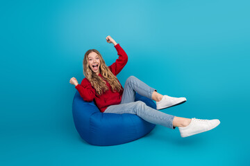 Adorable and cheerful young woman seated on a bean bag against a vibrant blue background, wearing...
