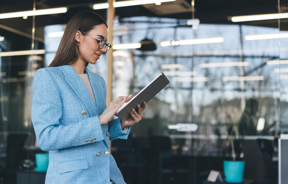 Young businesswoman in blue blazer uses digital tablet in office, representing fluid multitasking, modern autonomy and seamless engagement with smart technology.