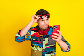 Young man with colorful shirt and red accessories looks at his phone against a bright yellow background