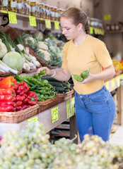 Young woman stands near the counter in a vegetable store and chooses fresh green peppers, she looks at them before weighing and buying.