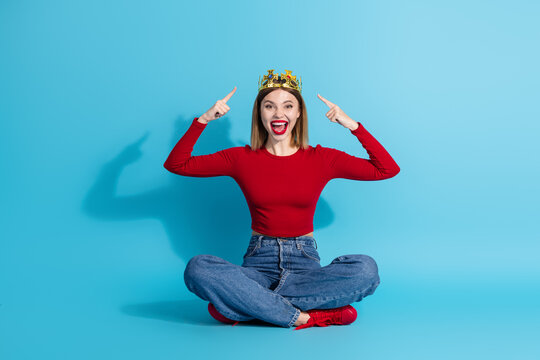 Lovely young woman wearing a crown seated cross-legged with a joyful expression against a vibrant blue background