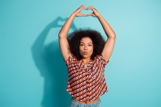 Fototapeta Young woman forms a heart with her arms against a blue background wearing a colorful patterned shirt