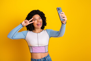 Smiling young woman making a peace gesture while taking a selfie in front of a vibrant yellow...