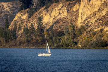 Scenic image of a white sailboat on a blue lake with rocky hills and trees in the background.