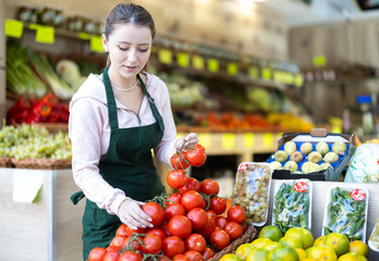 Young woman seller in apron puts fresh tomatoes on display at vegetable market