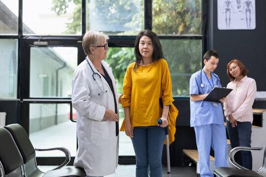 Medical expert helping Asian woman walk with mobility aid during routine hospital checkup. Specialist encouraging person to focus on injury recovery steps for independent movement.