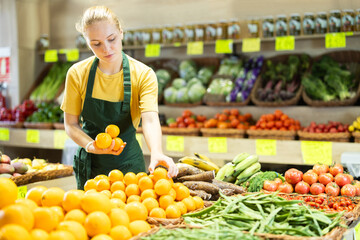 Teenage girl seller in apron puts fresh oranges on display in vegetable shop