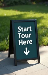Black framed sign on sidewalk says Start Tour With white arrow pointing down. Sign set against background of green grass, sunlight. Directional marker welcomes visitors to begin adventure exploration.
