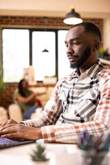 Obraz premium African american male manager typing on laptop, remotely replying to office emails and updating financial reports. Focused black man blogging online using digital device in brick wall apartment.