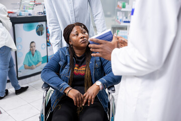 Black male pharmacist offers expert advice to man in wheelchair, providing assistance in choosing medication and vitamins for pharmaceutical care and wellbeing. Pharmacy shop service.