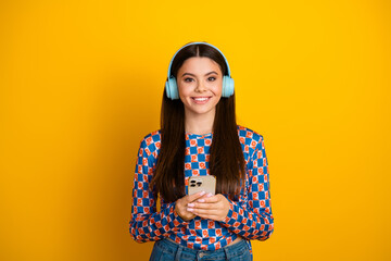 Young girl with blue headphones smiling while holding smartphone against bright yellow background for lifestyle fashion and shopping