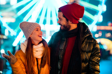 Cheerful couple enjoy a winter fair night with bright lights and warm smiles under festive...