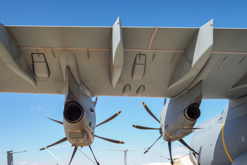 Underside view of military transport aircraft wing with twin turboprop engines and propeller blades against blue sky
