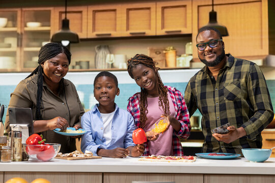 Portrait of cheerful parents cooking with kids to make homemade pizza together in a bright kitchen, spreading tomato sauce and layering pepperoni. Giving advice and tips to learn culinary hobby.