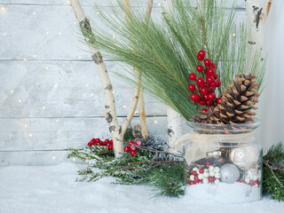 Christmas glass jar with ornaments and pinecones on snow, birch tree branches and festive holiday twinkle fairy lights, greenery and red berries on white wood plank rustic backdrop.