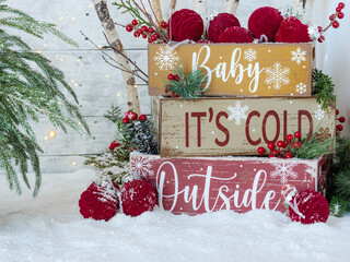 Christmas stacked crates on snow, holiday text Baby It’s Cold Outside, velvet maroon burgundy ornaments, birch and festive fairy lights, greenery and red berries on white wood plank rustic backdrop.